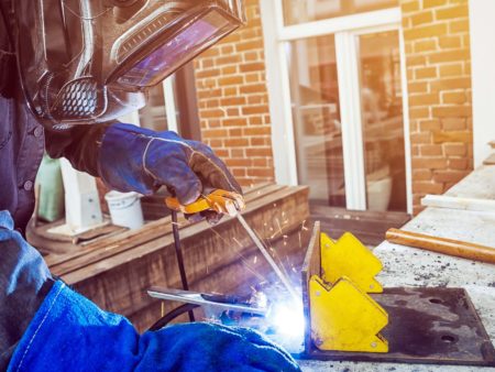 Man welder wearing a welding mask, building uniform and blue protective gloves brewing a metal welding machine on a street in a summer day, in the background an old brick building with a window and tools