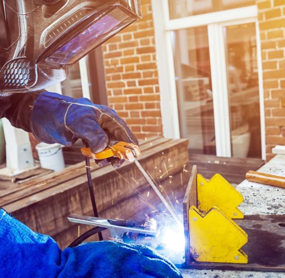 Man welder wearing a welding mask, building uniform and blue protective gloves brewing a metal welding machine on a street in a summer day, in the background an old brick building with a window and tools